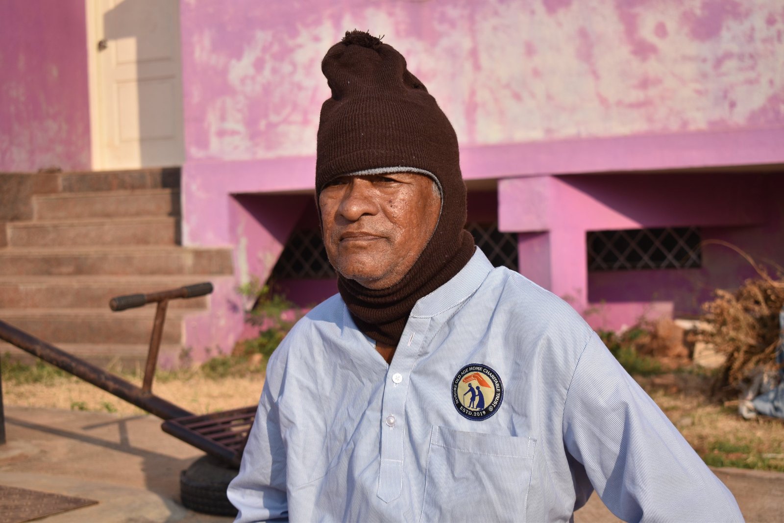 An elderly resident in a warm knitted cap and the home's uniform shirt, sitting outside in the winter sun
